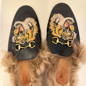 Gucci loafers med tigerbroderi och päls - Unika svarta loafers från Gucci med broderat tigerhuvud i gult, vitt och rött på ovansidan. Skorna har guldfärgade metallspännen och är fodrade med beige päls för extra statement. Tillverkade i skinn med platt sula och rund tå. Perfekt för dig som gillar att sticka ut.