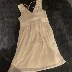 Champagnefärgad kort klänning från Vila - Champagnefärgad kort klänning från Vila i storlek S. Klänningen har v-ringad omlottfront, pärldetaljer på axlarna och ett brett midjeband som ger en snygg siluett. Materialet är lätt och luftigt, perfekt för varmare dagar. Axelbanden är smala och korsas i ryggen.