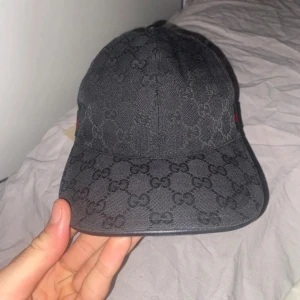  Gucci   - Snygg svart keps från Gucci med klassiskt GG-monogram över hela kepsen. Kepsen har böjd skärm och är tillverkad i bomullsmaterial. Perfekt accessoar för att lyfta din streetwear-look. 10/10