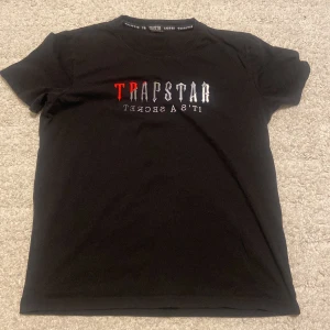 Svart Trapstar t-shirt med tryck - Svart t-shirt från Trapstar med stort broderat tryck på bröstet i rött och vitt där det står 'Trapstar It's a Secret'. Klassisk rund hals och korta ärmar. Materialet är mjuk bomull som känns skön mot huden. Perfekt för dig som gillar streetwear och vill sticka ut.