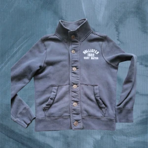 Mörkblå cardigan från Hollister - Snygg mörkblå cardigan jacka från Hollister med hög krage och knappar hela vägen framtill. Jackan har två fickor och tryckt logga på bröstet. Mjuk insida i bomullsmix, perfekt för chill dagar. Klassisk och enkel design som funkar till det mesta.