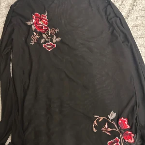 Svart meshblus med broderade rosor - Svart transparent meshblus från KappAhl i storlek M. Blusen har lång ärm och är dekorerad med broderade röda rosor och blad framtill och nedtill. Perfekt för dig som gillar detaljer och vill sticka ut med blommigt broderi.