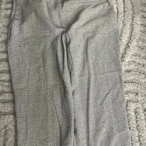 Grå rutiga kostymbyxor från H&M - Snygga grå kostymbyxor från H&M i storlek 38 med klassiskt rutigt mönster. Byxorna har rak passform, dragkedja och knapp framtill. Perfekta för en stilren look och passar dig som gillar tidlös design.