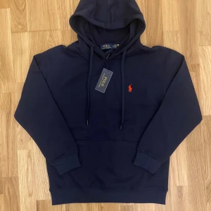 Marinblå hoodie från Polo Ralph Lauren - Snygg marinblå hoodie från Polo Ralph Lauren med klassisk röd broderad logga på bröstet. Tröjan har huva med snörning, ribbade muddar och en stor magficka. Tillverkad i mjuk bomull som är skön att ha på sig. Perfekt för dig som gillar stilrena och ikoniska plagg. Lite kort vid armarna.
