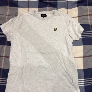 Ljusgrå t-shirt från Lyle & Scott - Snygg ljusgrå t-shirt från Lyle & Scott med klassisk rund hals och korta ärmar. På bröstet sitter den ikoniska gula fågeln som broderad detalj. Perfekt basic-plagg i mjuk bomull som funkar till jeans eller shorts.