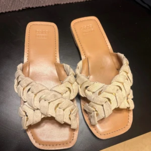 Beige flätade sandaler från ASOS - Snygga sandaler från SOSO med flätad rem i beige tyg och platt sula. Sandalerna har fyrkantig tå och en stilren design som passar perfekt till varma dagar. Materialet är mjukt tyg och syntet, med detaljer i guld på remmarna.