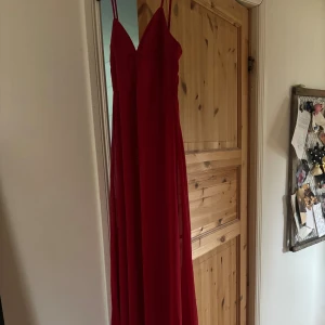 Röd långklänning med smala axelband - Snygg röd långklänning med smala axelband och v-ringad urringning. Klänningen har en elegant och flowy passform, dragkedja i ryggen och är ärmlös. Perfekt för bal eller student. Materialet känns lätt och följsamt.