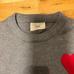 Grå stickad tröja från Ami med hjärta - Säljer en grå stickad tröja från Ami med rund hals och ribbade muddar. På bröstet finns ett rött hjärta med bokstaven A, vilket ger en unik och snygg detalj. Perfekt för dig som gillar stilrena plagg med en twist.