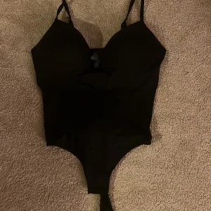 Svart bodysuit från H&M, storlek L - Säljer en svart bodysuit från H&M i storlek L. Modellen har smala axelband och en djup v-ringning framtill. Materialet är en mix av bomull och polyester med elastan för stretch. Perfekt att styla med jeans eller kjol för en clean look.