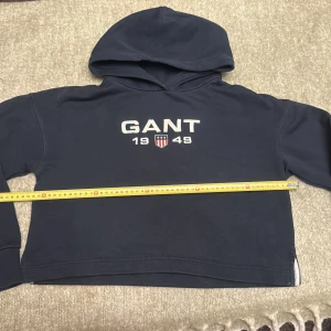 Marinblå hoodie från GANT - Snygg marinblå hoodie från GANT med broderad logga och texten '1949' framtill. Tröjan har huva, långa ärmar med ribbade muddar och är tillverkad i mjuk bomullsmix. Perfekt för en chill och sportig look.