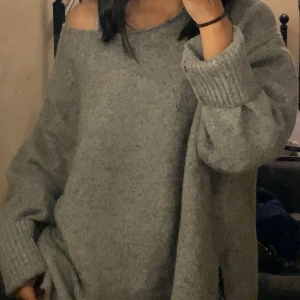 Grå offshoulder stickad tröja H&M - Säljer en oversized grå stickad tröja från H&M i storlek L. Tröjan har en snygg offshoulder-design och breda ribbade muddar vid ärmslut. Perfekt för dig som gillar chill och avslappnad stil med lite edge.