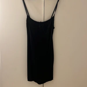 Svart kort sidenklänning med rosett - Svart kort sidenklänning med tunna axelband och liten rosett framtill. Klänningen har spetsdetaljer längs urringningen och är tillverkad i ett glansigt, mjukt material som ger en elegant känsla. Perfekt längd🥰