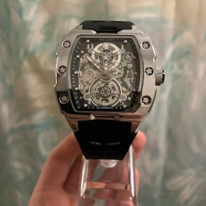 Megir automatisk klocka  - Säljer en snygg automatisk klocka från Megir med genomskinlig urtavla där du ser mekaniken. Klockan har en robust silverfärgad boett, svarta detaljer och svart armband i syntetmaterial. Rektangulär form med coola skruvdetaljer och tydliga siffror.                                            ✅Helt Ny✨.                                                                                    ✅Frakt inom 24 timmar📦.                                                      ✅Kommer med Låda 🗃️.