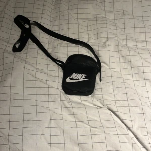 Svart axelväska från Nike - Snygg svart axelväska från Nike med justerbar rem och vit logga framtill. Väskan har dragkedja och ett vinrött innerfoder med flera små fack. Perfekt storlek för mobil, plånbok och andra småprylar. Enkel och stilren design som funkar till det mesta.