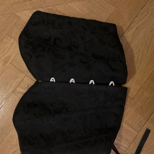 Svart korsett med snörning - Säljer en svart korsett med snyggt mönstrad yta och glittriga detaljer. Den har metallknäppning framtill och snörning bak för perfekt passform. Korsetten har en markerad midja och är tillverkad i ett glansigt syntetmaterial. Perfekt för att lyfta vilken outfit som helst.