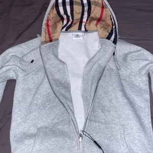Grå Burberry hoodie - Säljer en grå hoodiejacka från Burberry med klassiskt rutigt foder i huvan. Jackan har dragkedja den är helt autentisk och ny. Skickas inom 2 dagar