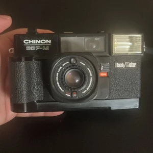 Chinon 35F-M - Chinon 35F-M är en klassisk analog kamera med inbyggd blixt och motorframdragning. Utrustad med 38mm f/2.8 Chinonex Color-lins. Kameran ser ut att vara i gott skick med mindre bruksspår, och filmfacket är rent. Perfekt för dig som vill börja med analog fotografering.