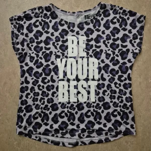 T-shirt - Cool t-shirt med leopardmönster i svart och mörklila på ljusgrå botten. Stor vit text 'BE YOUR BEST' framtill. Kortärmad och tillverkad i mjuk polyester, perfekt för dig som gillar att sticka ut. Rund hals och loose passform.