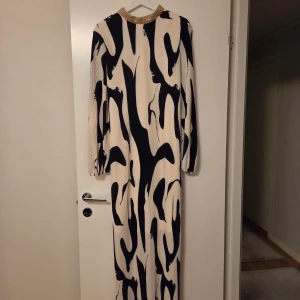 Långklänning med svartvitt mönster & guldkrage - Säljer en maxiklänning med svart och vitt abstrakt mönster, långa ärmar och snygg guldglittrig krage. Klänningen har plisserat tyg och ett knytband i midjan för extra passform. Perfekt för dig som vill sticka ut med en cool och elegant look.