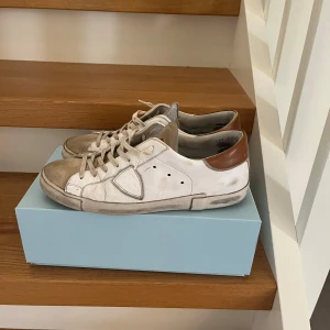 Philippe Model vita sneakers i skinn - Snygga vita sneakers från Philippe Model med beige mockadetaljer på tå och plös samt brunt skinn vid hälen. Klassisk låg modell med snörning och rund tå. Sulan har tydlig logga undertill. Perfekta för en stilren och avslappnad look. Lite slitna i hälen men fortfarande mycket bra skick. 