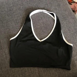 Svart croppad linnetopp med vit kant - En svart croppad topp med vita kanter och djup v-ringning. Toppen har halterneck. Materialet är silkit och stretchigt, den är S men passar M oxå