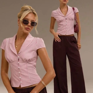 Rosa och vit randig blus med knappar - Säljer en kortärmad blus med rosa och vita ränder, figurnära passform och snygg krage. Blusen har knappar framtill och lätt markerad midja. Perfekt till vida byxor eller jeans för en trendig look. 🥰