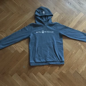 Blå hoodie från Sail Racing - Blå hoodie från Sail Racing med vit logga på bröstet och huvan. Tröjan har en klassisk känguruficka framtill och är tillverkad i mjuk bomull med fleece på insidan. Långärmad och normal passform, perfekt för chill dagar.