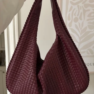 Vinröd flätad axelväska Design by si - Säljer en vinröd axelväska med snygg flätad design. Från design by si och i nyskick.