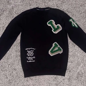 Svart Louis Vuitton sweatshirt med patchar - Svart sweatshirt från Louis Vuitton med gröna och vita patchar i form av bokstäver på bröstet och ärmen. Tröjan har rund hals, långa ärmar och vit textbroderi på framsidan. Materialet är mjuk bomull och passformen är normal.