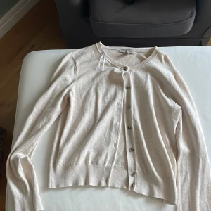 Beige kofta från H&M med knappar - Snygg beige kofta från H&M i storlek S. Koftan har rund halsringning, långa ärmar och knappar framtill. Materialet känns mjukt och lätt, perfekt för lager-på-lager. Ribbstickade muddar vid ärmslut och nederkant ger en clean look.