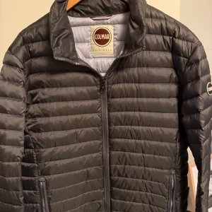 Svart pufferjacka från Colmar - Snygg svart pufferjacka från Colmar med horisontella quiltade paneler och dragkedja framtill. Jackan har ståkrage, två sidofickor med dragkedja och Colmar-logga på ärmen. Perfekt för kyliga dagar och har en clean, modern look.