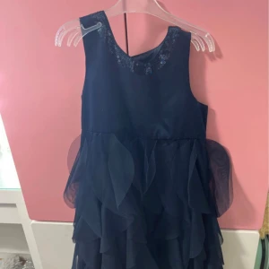 Marinblå klänning med paljetter - Snygg marinblå klänning med ärmlös design och rund halsringning dekorerad med paljetter. Kjolen har flera lager med volanger i tunt tyg som ger en cool och festlig look. Stängs med knappar i ryggen. Perfekt för dig som vill sticka ut.