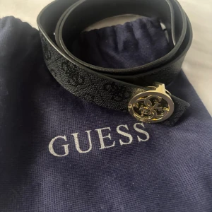 Svart bälte med guldspänne från Guess - Snyggt svart bälte från Guess med diskret logomönster och ett rundat guldigt spänne med Guess-logga. Bältet är smalt och har en stilren design som lyfter vilken outfit som helst. Materialet är skinn och bältet kommer i originalpåse.