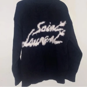 Svart stickad Saint Laurent tröja - Säljer en svart stickad tröja från Saint Laurent med vit, fluffig logotyp framtill. Tröjan har rund hals och långa ärmar, perfekt för dig som gillar statement-plagg med lyxig vibe. Passar dig som vill sticka ut med designerstil.
