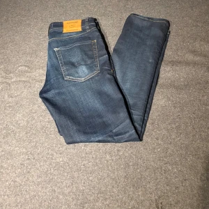 Jack & Jones Slim Straight jeans blå - Snygga mörkblå jeans från Jack & Jones i modellen Slim Straight Tim. Klassisk femficksdesign, kontrastsömmar och knappgylf. Passformen är slim med raka ben, perfekt för dig som gillar en stilren look. Storlek 29/30.
