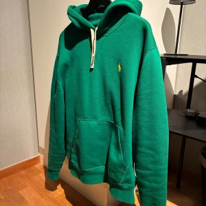 Grön hoodie från Polo Ralph Lauren - Snygg grön hoodie från Polo Ralph Lauren med klassisk gul logga på bröstet. Tröjan har huva med vita snören och en stor känguruficka framtill. Perfekt för dig som gillar en avslappnad och stilren look.