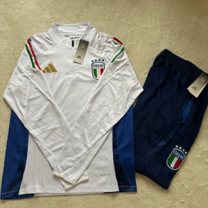 Italien långärmad fotbollströja Adidas - Säljer en officiell långärmad Italien fotbollströja från Adidas i vit polyester med blå detaljer på sidorna. Tröjan har broderat landslagsmärke, guldigt Adidas-logo och ränder i grönt och rött på axlarna. Halv dragkedja framtill och Aeroready-material för extra komfort.