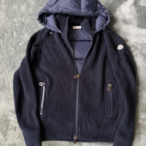 Mörkblå stickad cardigan jacka Moncler - Snygg mörkblå stickad cardigan från Moncler med dunfylld huva och insida. Har dragkedjeförsedda fickor, ribbade muddar och Moncler-logga med NFC som leder till deras hemsida. Perfekt för en vanlig dag i skolan – varm, exklusiv och sportig.