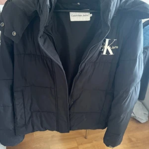 Svart Calvin Klein Jacka  - Säljer min fina vinterjacka från Calvin Klein i storlek XL. Jackan är varm, bekväm och har en stilren design. Den har dragkedja, huva och fickor framtill. Jackan är i mycket bra skick och använd endast 1-2 gånger.