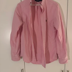 Oxfordskjorta från Polo Ralph Lauren - Snygg rosa skjorta från Polo Ralph Lauren i slim fit med klassisk button-down krage och broderad logga på bröstet. Skjortan har långa ärmar, knappar framtill och är gjord i mjuk bomull. Perfekt för dig som gillar stilrena och färgglada plagg.