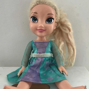 Frost docka Elsa - Säljer en dockklänning från JAKKS i turkos och lila med glittrigt bälte och transparenta ärmar. Klänningen har satinkänsla och fina tryckta detaljer framtill. Perfekt för dig som gillar sagolika och färgglada outfits till dockan.