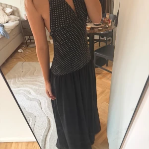 Svart prickig långklänning med halterneck - Svart långklänning med vita prickar upptill och helsvart kjol. Snygg halterneck med knyt i nacken och djup urringning framtill. Öppen rygg och figurnära passform upptill, kjolen är luftig och faller snyggt. Nyskick, endast använd en gång 