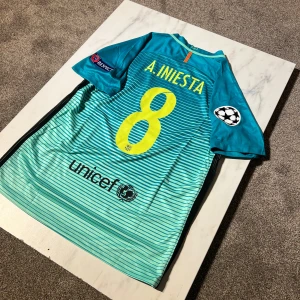 Barcelona Iniesta bortatröja Nike M - Säljer en officiell FC Barcelona bortatröja från Nike med Iniesta och nummer 8 på ryggen. Tröjan är turkosblå med tunna horisontella ränder, Champions League-patchar på ärmarna och klubbmärke på bröstet. Materialet är lätt och andas, perfekt för match eller samling.