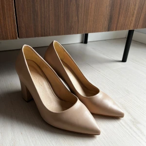 Beige pumps med klack från London & London - Snygga beige pumps från London & London med spetsig tå och stabil blockklack. Skorna är tillverkade i slätt skinnmaterial och har en klassisk, stilren design som passar perfekt till dig som gillar en clean och elegant look.