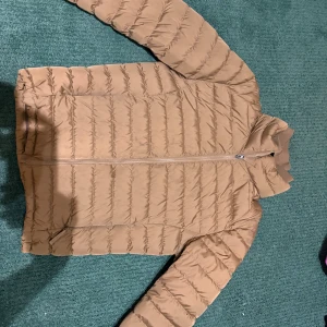 Beige pufferjacka med dragkedja - Säljer en beige pufferjacka med horisontella quiltade sömmar och hel dragkedja framtill. Jackan har huva och långärmade armar, perfekt för kyliga dagar. Stilren och enkel design som passar till många olika outfits.