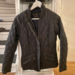 Svart quiltad jacka från Barbour - Quiltad svart jacka från Barbour med klassisk krage, dragkedja och tryckknappar framtill. Jackan har två fickor med knappstängning och snyggt rutmönster över hela ytan. Perfekt för dig som gillar stilren och tidlös design. I gott skick 