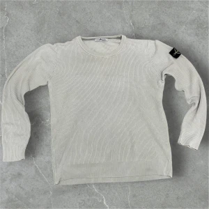 Stickad Stone Island Tröja - Säljer en ljusgrå ribbad tröja från Stone Island med klassisk rund hals och ikonisk patch på ärmen. Tröjan är tillverkad i 100% bomull och har lång ärm. Perfekt för dig som gillar stilrena och exklusiva plagg.