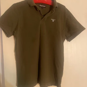 Olivgrön pikétröja från Barbour - Snygg olivgrön pikétröja från Barbour i klassisk modell med krage och korta ärmar. Tröjan har Barbour-logga broderad på bröstet och är gjord i mjuk bomull. Perfekt för dig som gillar stilrena och tidlösa plagg.