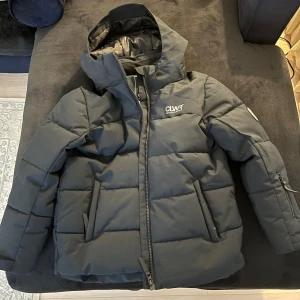 Svart pufferjacka från CLWR - Svart pufferjacka från CLWR med huva och dragkedja framtill. Jackan har två sidofickor med dragkedja och CLWR-logga på bröstet. Perfekt för kalla dagar med sin tjocka och quiltade design. Nypris 1500 kr!