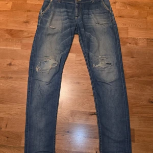 Blå slitna jeans från Dondup - Säljer ett par blå jeans från Dondup med snygga slitningar och hål på benen för en cool, avslappnad look. Modellen har klassisk femficksdesign, normal passform och raka ben. Jeansen är tillverkade i jeansmaterial och har detaljerade sömmar. Storlek 14 år, passar dig som är runt 175cm lång. Skriv vid minsta fråga och våga buda!!